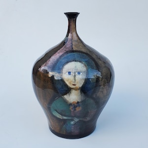 May include: A ceramic vase with a dark brown glaze and a painted portrait of a woman with blue eyes. The woman is wearing a green dress and has a floral pattern on her chest.