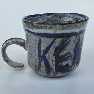 May include: A handmade ceramic mug with a gray speckled finish and a curved handle. The mug features blue brushstrokes and the word "KE" in blue. The rim has a dark green and white band.