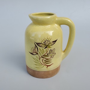 May include: A yellow ceramic pitcher with a brown handle and a floral design in brown and green. The pitcher has a crackled glaze and a brown base.
