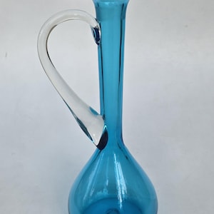 May include: A tall, translucent blue glass pitcher with a bulbous base, a slender neck, and a wide, flared rim. The vessel features a single, clear, curved glass handle attached to the side, extending from the neck down to the rounded body.