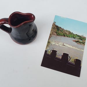 May include: A small, dark-colored ceramic pitcher with a handle and a red rim. Next to it is a postcard featuring a river scene with a blue sky and a row of pitcher silhouettes.