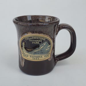 May include: A brown ceramic mug with a handle, featuring an oval emblem with the text "Yellowstone National Park" and "Old Faithful Inn 1904". The mug has a textured, speckled finish.