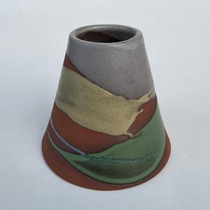 May include: A conical ceramic vessel featuring a matte finish with abstract color blocking in shades of terracotta, sage green, muted yellow, and grey. The object has a wide circular base that tapers upward to a smooth, open circular rim.
