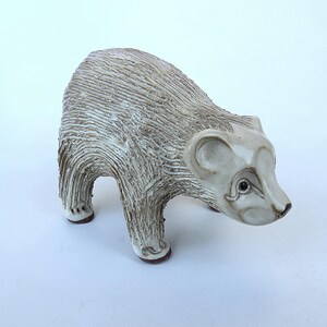 May include: A ceramic bear figurine featuring a textured, grooved surface in shades of off-white and light brown. The bear is depicted in a standing position with a smooth, glazed face, rounded ears, and dark, defined eyes, showcasing a handcrafted aesthetic.