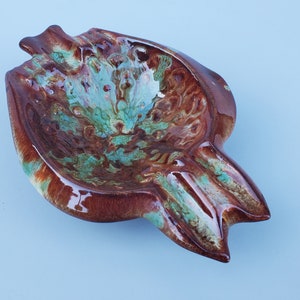 May include: A brown and green ceramic ashtray with a unique, abstract design. The ashtray is shaped like a bird with a wide, shallow bowl.