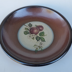 May include: A round, brown ceramic plate with a glossy finish. The center features a painted design of two red apples, green leaves, and white blossoms on a light background. The rim is dark brown.