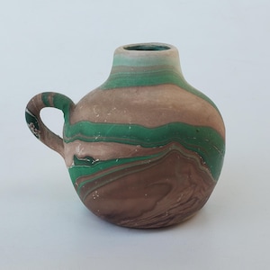 May include: A small, handcrafted ceramic vase with a handle. The vase has a distinctive swirl pattern in brown, green, and tan. The opening is light green. This is a decorative item.