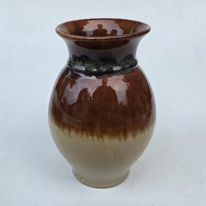 May include: A handcrafted ceramic vase with a glossy finish. The vase features a gradient of colors, transitioning from a light beige base to a rich brown at the top. The rim is a darker shade of brown.