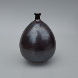 May include: A dark, pear-shaped ceramic vase with a narrow neck. The vase has a speckled, metallic finish in shades of dark gray and burgundy. The base is a lighter color, and the vase is set against a neutral background.