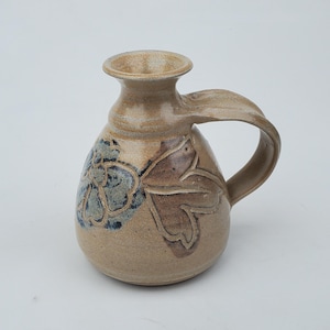 May include: A small, beige ceramic jug with a brown and blue floral design. The jug has a handle and a rounded body.