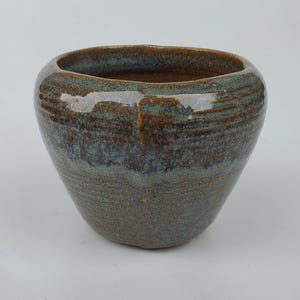 May include: A handcrafted ceramic bowl with a wide opening and a rounded base. The bowl features a textured surface with a blend of earthy tones, including brown, green, and blue hues. The interior is a darker shade of brown.