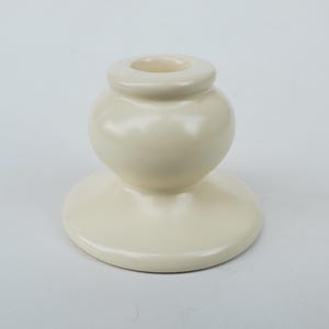 May include: Off-white ceramic candle holder with a rounded body and a circular base. The top features a small, recessed area for holding a candle. The smooth, glossy finish gives it a clean, modern look. This decorative piece is suitable for any room.