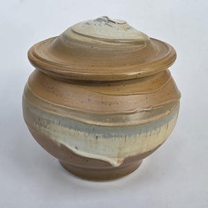 May include: A round ceramic jar with a matching lid, featuring a speckled tan and brown glaze with a horizontal band of muted blue and cream drip patterns around the center. The vessel has a wide, curved body and a slightly raised, rounded lid.