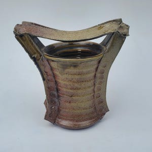 May include: A brown ceramic vase with a unique handle that wraps around the top. The vase has a textured surface and a slightly flared opening.