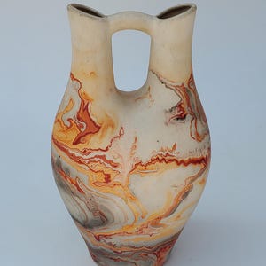 May include: A ceramic vase with a distinctive double-spout design and a central handle. The vase has a marbled pattern in shades of orange, red, grey, and cream. The vase is a decorative item, with a striking visual effect.