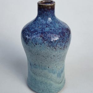 May include: A ceramic vase with a narrow neck and a wide body, featuring a reactive glaze that transitions from a dark, deep blue at the top to a lighter, mottled sky blue and pale grey at the base, with a smooth, glossy finish throughout.
