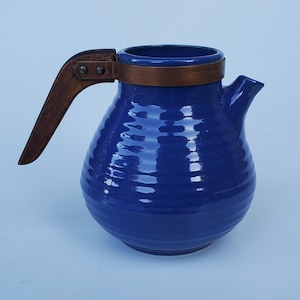 May include: A cobalt blue ceramic pitcher with a wooden handle and copper band. The pitcher has a rounded body with horizontal ridges and a spout. The handle is attached with metal rivets. The pitcher is likely used for serving beverages.