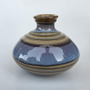 May include: A round ceramic vase with a wide, bulbous body and a narrow neck. The glaze features a blend of deep blue, brown, and tan tones with horizontal bands circling the body. The surface has a glossy finish with a reflective quality.