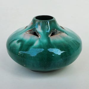 May include: A handcrafted, teal-green ceramic vase with a rounded body and a narrow neck. The vase features three triangular openings around the top and a glossy finish with subtle color variations. A unique decorative piece.