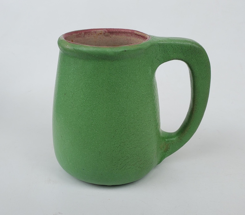 May include: A green ceramic mug with a rounded body and a large handle. The interior rim is a reddish-brown color. The mug has a textured surface and is set against a white background. This handmade mug is perfect for coffee or tea.