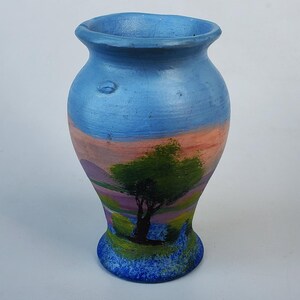 May include: A small, blue ceramic vase with a painted landscape scene. The vase has a rounded body and a flared lip. The scene features a green tree, pink and purple hills, and a blue sky.