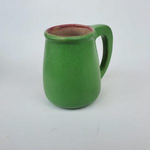 May include: A small, green ceramic mug with a rounded body and a handle. The interior rim is a reddish-brown color. The mug has a textured surface and is set against a white background. This handmade mug is perfect for coffee or tea.
