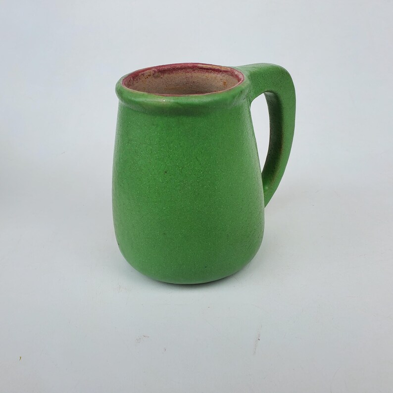 May include: A small, green ceramic mug with a rounded body and a handle. The interior rim is a reddish-brown color. The mug has a textured surface and is set against a white background. This handmade mug is perfect for coffee or tea.