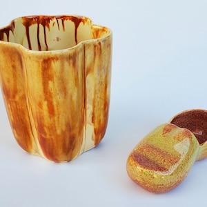 May include: A ceramic container and lid set. The container is a tall, fluted shape with a cream-coloured base and brown glaze that drips down the sides. The lid is a rounded, oval shape with a similar glaze. The set is on a white background.