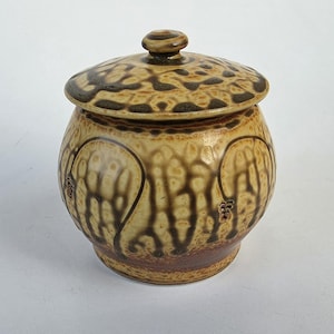 May include: A ceramic jar with a lid. The jar is round with a bulbous base and a lid with a small knob. The jar is tan with brown patterns and accents. The lid has a darker brown pattern. The jar is likely used for storage.
