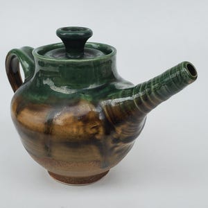 May include: A handcrafted ceramic teapot with a dark green lid and handle. The body of the teapot is a mix of brown and green hues, with a long spout. The teapot is designed for brewing tea.
