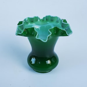 May include: A dark green glass vase with a ruffled rim. The vase has a bulbous base and a flared neck. The rim is a lighter shade of green. The vase is approximately 7 inches tall.