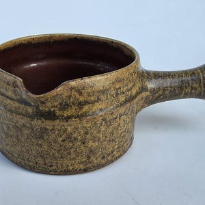 May include: A handmade, ceramic saucepan with a handle. The exterior is a mottled brown and tan, with a dark brown interior. The spout is on the left side. This rustic kitchenware is ideal for cooking and serving.