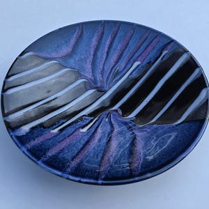 May include: A round ceramic bowl featuring a glossy glaze with a deep blue and black base. The surface is decorated with radiating, textured white and purple streaks that extend from the center toward the outer rim, creating a distinct pattern.