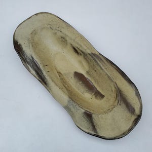 May include: A ceramic serving dish with a light brown glaze and dark brown streaks. The dish has an oval shape and a slightly raised rim.