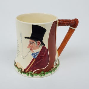 May include: A ceramic mug with a brown handle shaped like a pipe. The mug features a hand-painted illustration of a man in a top hat and a red jacket, smoking a pipe. The mug has a white background with green leaves around the bottom.