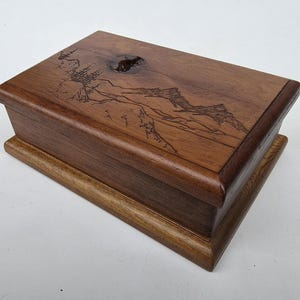 Peut inclure: Une boîte rectangulaire en bois avec un paysage de montagne gravé sur le couvercle. La boîte est en bois brun foncé avec une base brun clair. Le couvercle a des bords arrondis et un nœud dans le bois.