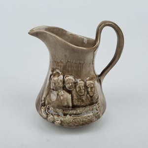 May include: A brown ceramic pitcher with a handle and spout. The pitcher features a relief of four faces and the words "MOUNT RUSHMORE" and "BLACK HILLS, S". A decorative souvenir item.