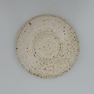 May include: A white ceramic saucer with a speckled brown glaze. The saucer has a slightly raised rim and a smooth, rounded bottom.