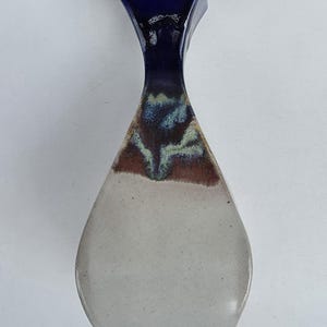 May include: A ceramic vase with a unique shape and color gradient. The top is a deep blue, transitioning to brown and green in the middle, and fading to a light gray at the base. The vase has a square top and a teardrop-shaped body.