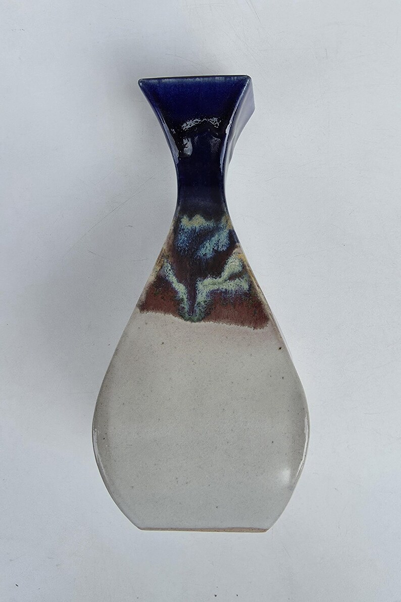 May include: A ceramic vase with a unique shape and color gradient. The top is a deep blue, transitioning to brown and green in the middle, and fading to a light gray at the base. The vase has a square top and a teardrop-shaped body.