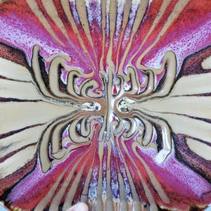 May include: A decorative ceramic plate with a unique, abstract design. The plate features a radial pattern with shades of red, brown, beige, and black. The design has a flowing, organic quality, with a central, symmetrical motif.