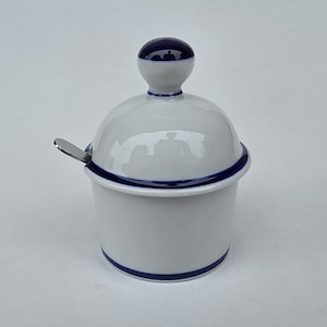 May include: White ceramic sugar bowl with a blue rim and a matching blue-topped lid. A small stainless steel spoon rests on the side. The bowl is round and has a simple, elegant design.