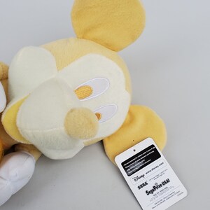 Puede incluir: Un juguete de peluche Mickey Mouse amarillo con detalles blancos. El juguete est&aacute; tumbado de lado con la cabeza girada hacia la derecha. El juguete tiene una etiqueta pegada a la oreja derecha que dice "Disney Sega Prize USA!" y "Solo para canje de premios".