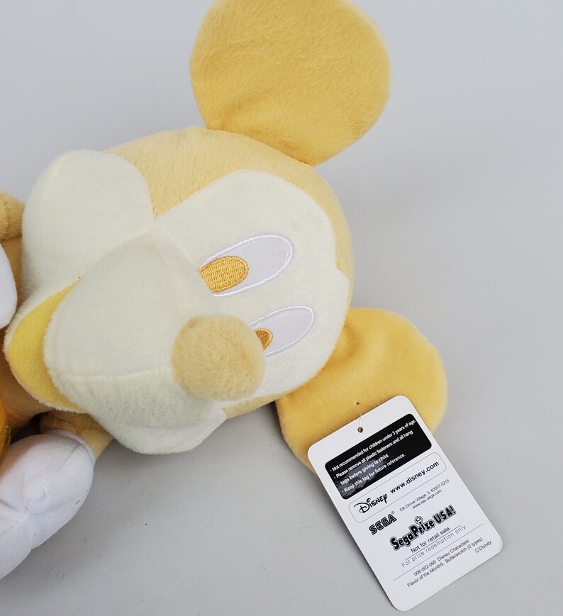 Puede incluir: Un juguete de peluche Mickey Mouse amarillo con detalles blancos. El juguete est&aacute; tumbado de lado con la cabeza girada hacia la derecha. El juguete tiene una etiqueta pegada a la oreja derecha que dice "Disney Sega Prize USA!" y "Solo para canje de premios".