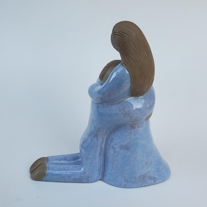 May include: A ceramic sculpture of a kneeling figure in a light blue dress, embracing another figure. The sculpture has brown hair and a serene expression. The piece is a unique art object.