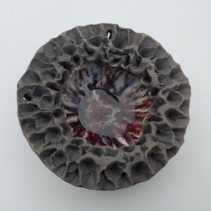 May include: A ceramic bowl with a textured, dark gray exterior and a deep, red and purple glazed interior. The bowl has a unique, organic shape.