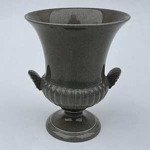 May include: A dark gray urn-shaped vase with a flared top and a pedestal base. The vase features two decorative handles and a ribbed design around the midsection. The overall height is approximately 12 inches.