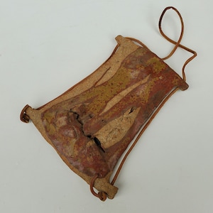May include: A small, brown leather pouch with a tan and red patterned design. The pouch has a leather strap that can be used to hang it.