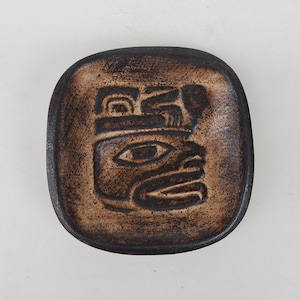 May include: A square, brown ceramic dish with a raised, stylized face design. The dish has a dark brown rim and a lighter brown interior, with the face design in a darker shade. The design appears to be an abstract representation of a face.