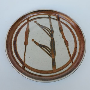 May include: A white ceramic plate with a brown rim and a stylized design of brown reeds on the surface.
